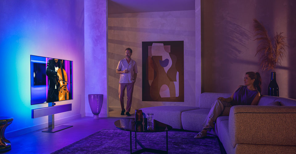 Vīrietis un sieviete stilīgā viesistabā skatās Philips Ambilight televizoru, kurā tiek rādīts čella priek&scaron;nesums