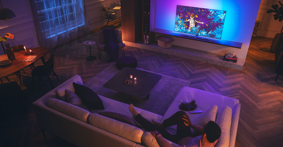 Vīrietis atpū&scaron;as uz dīvāna vāji apgaismotā viesistabā, skatoties Philips Ambilight televizoru