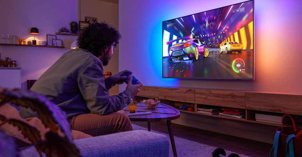 Cilvēks sēž uz dīvāna un spēlē sacīk&scaron;u videospēli Philips Ambilight televizorā